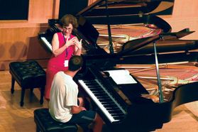student sits at piano while instructor stands and speaks next to him